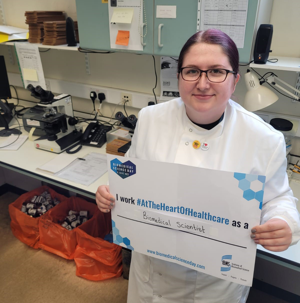 Elisha Hughes in a lab holding a BMS certificate noting that they work as a biomedical scientist