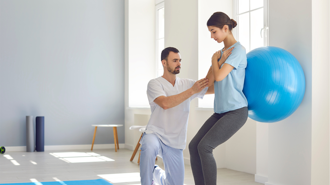 Female leaning on an exercise ball against a wall, with a male helping her