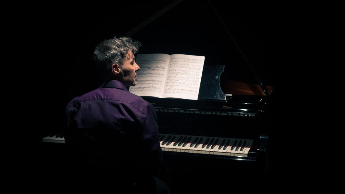 Dun mewn crys piws a gwallt byr yn llwydo tn edrych i'r dde. Rhydym yn ei weld mewn profil o'r cefn, ac yn gweld allweddellau'r piano, ond mae gweddill y llun yn ddu.t with short gerying hair plays piano and looks to the right, so we see his face in profile from the back, we see him and the piano keys the rest is black.