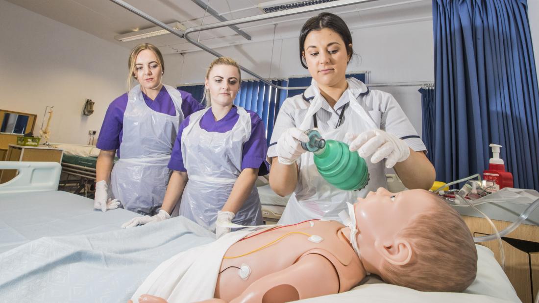 Dwy myfyrwraig nyrsio yn gwylio arddangosiad wrth ochr gwely  gyda dymi yn y gwely.ents at a bedside watching someone with a dummy