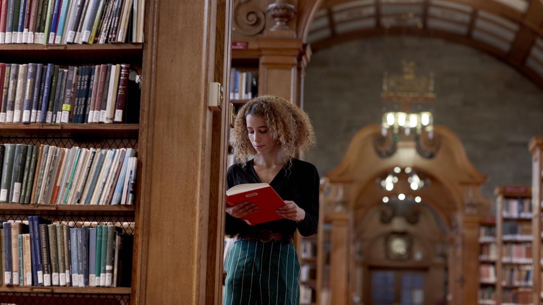 Student reading a book in Shankland Library leaning on book shelf. 