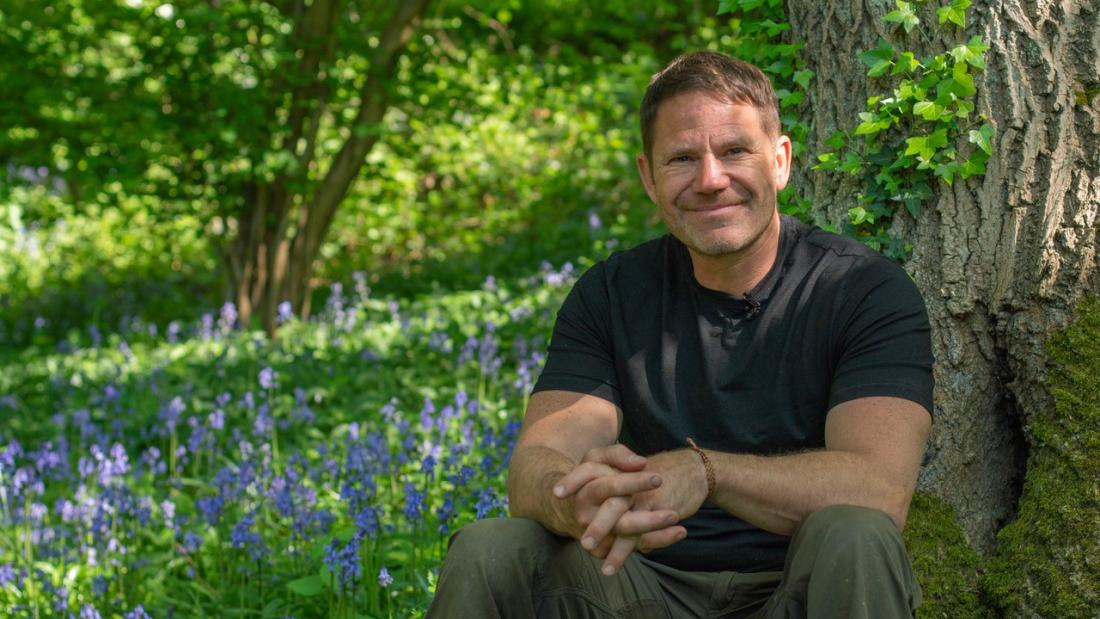 Eisteddai Steve Backshall tu allan, mae clychau'r gog mewn blodau o'i gwmpas a choed yn y cefndir.