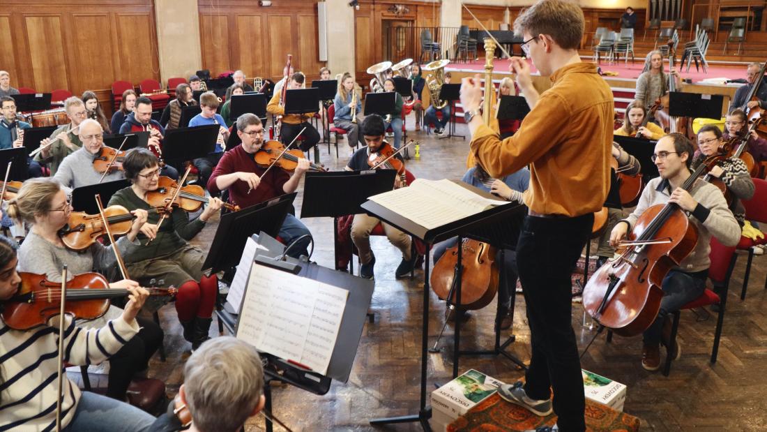  A young man in mustard coloured shirt conducts a n orchestra of adults and young people