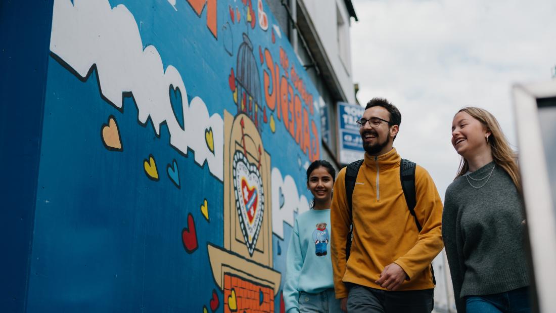 Three students walking in Bangor High Street next to wall mural