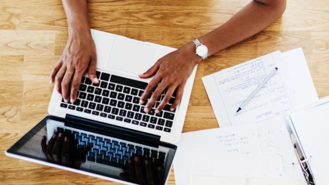 Hands typing on a laptop. Next to the laptop are a file, pen and notebook.