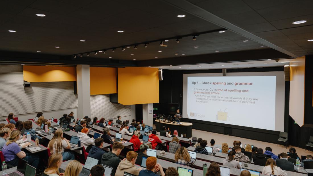 A view of the lecture theatre from the back of the room looking down on a room full of students looking at the projector screen at the front of the room.