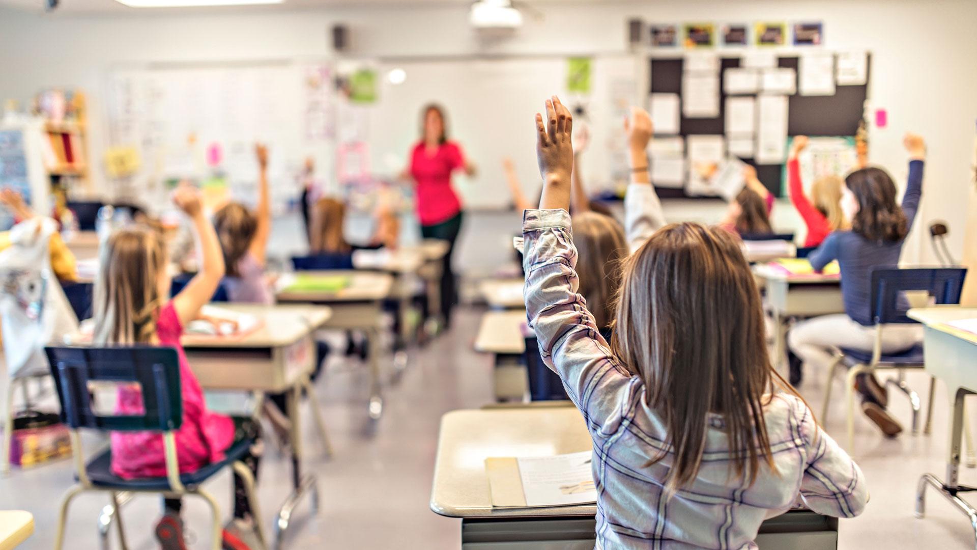 primary children in classroom holding up hands to answer a question