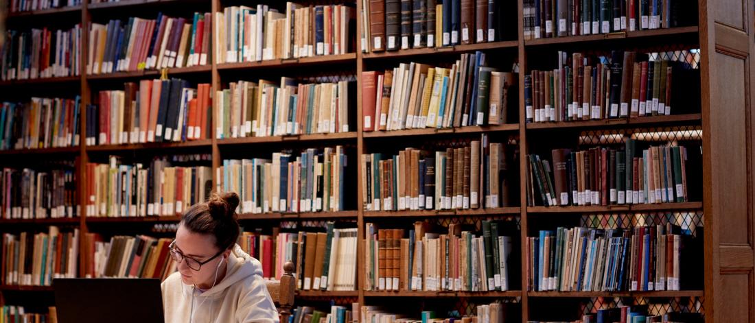 Student sitting in the library with a laptop and books on shelves in the background