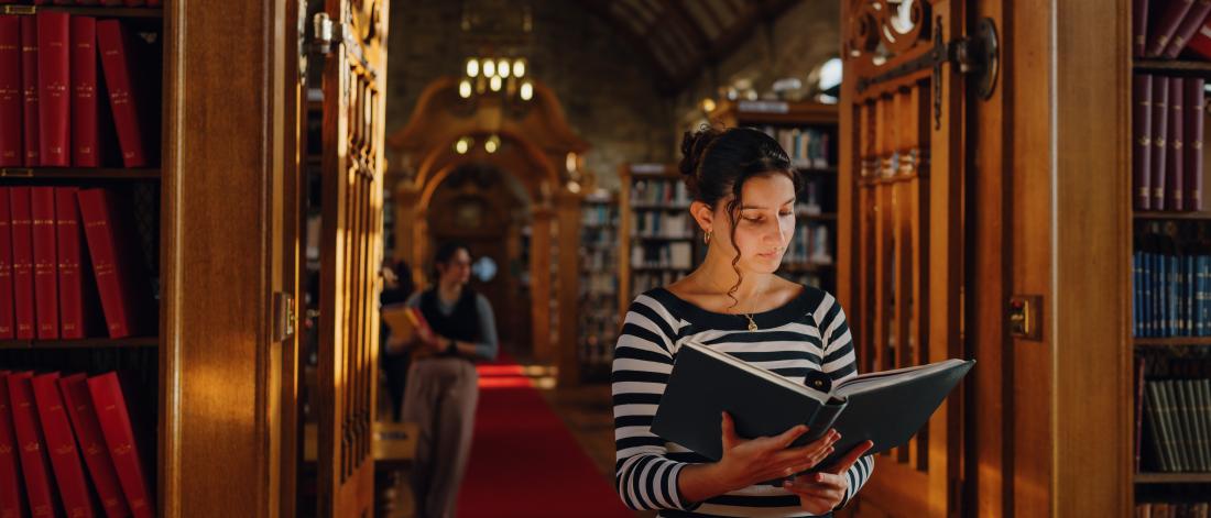 A student stands in the central aisle of Shankland Library reading a book, surrounded by bookshelves with warm light filtering through high, arched windows. The vaulted ceiling and chandeliers add a sense of quiet grandeur.