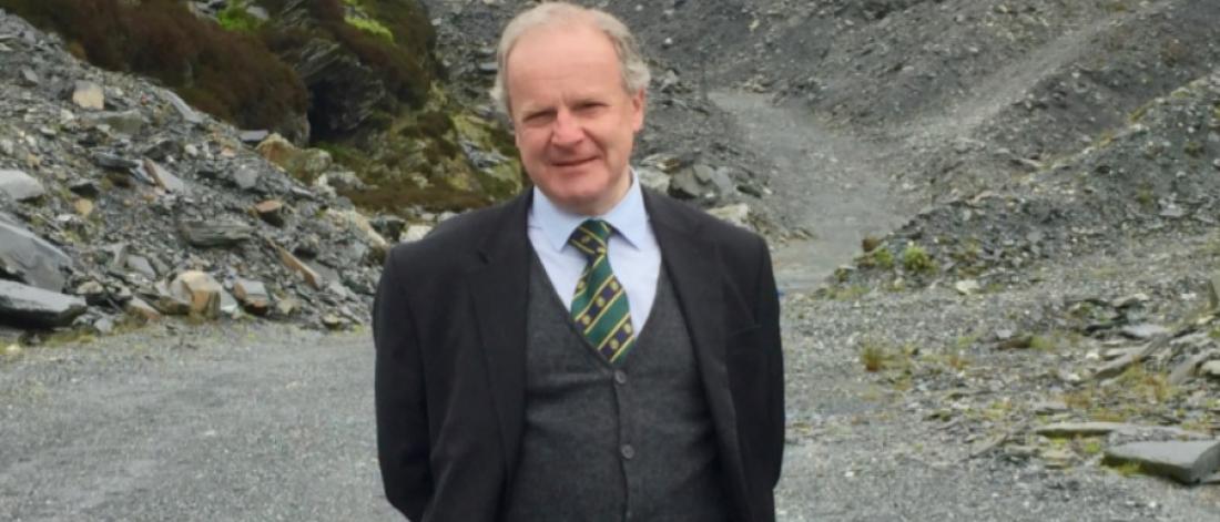 A photograph of David Gwyn smiling, behind him is an example of the slate landscape, with a path that is flanked by slate slag heaps 