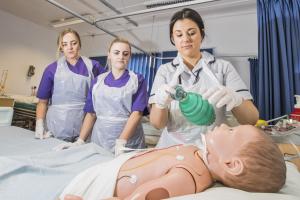 Dwy myfyrwraig nyrsio yn gwylio arddangosiad wrth ochr gwely  gyda dymi yn y gwely.ents at a bedside watching someone with a dummy