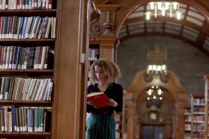 Student reading a book in Shankland Library leaning on book shelf. 