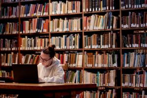 Student sitting in the library with a laptop and books on shelves in the background