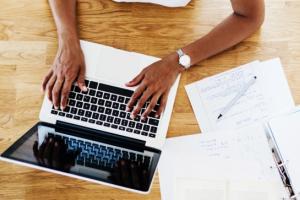 Hands typing on a laptop. Next to the laptop are a file, pen and notebook.