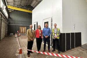 Three men and one woman standing in front of a small, white, block shaped building inside a large building