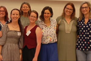 7 women stand in a row smiling. There is no visible background and all are wearing semi-formal conference clothes