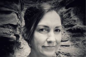 A black and white headshot of a woman with long dark hair, standing in front of a stone wall