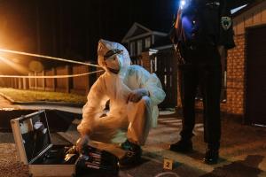 A forensic investigator in a white suit meticulously examines evidence at a dimly lit, cordoned-off crime scene with tools and police tape in the background.