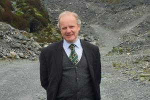 A photograph of David Gwyn smiling, behind him is an example of the slate landscape, with a path that is flanked by slate slag heaps 