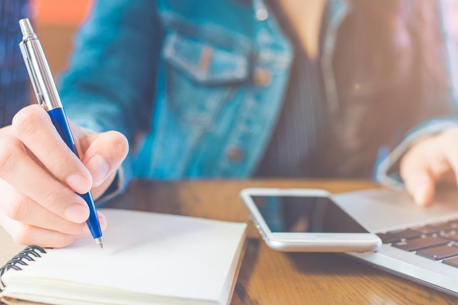 Close-up of student writing in notepad with mobile phone and laptop on desk