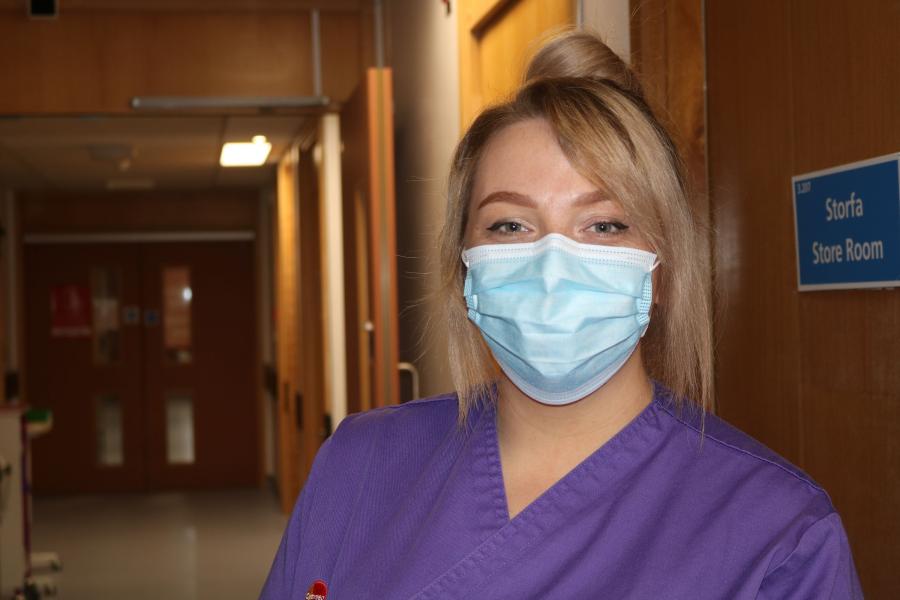 A student nurse wearing  purple uniform and a mask looks into the camera