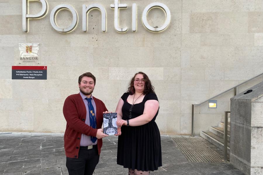 Dau fyfyriwr o Brifysgol Bangor yn dangos eu cyhoeddiad cyntaf y tu allan i adeilad Pontionts show the book they have published while standing in front f the external of the Pontio building