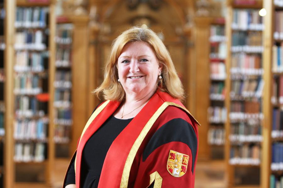 Woman with shoulder length  blonde hair smiles at the camera. In the background are the out of focus bookshelves of Bangor University library.