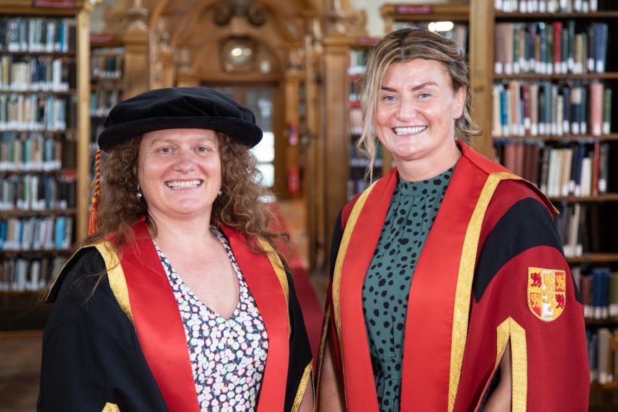 Two women stand side by side, one in Balck academic gown and one in a red gown.