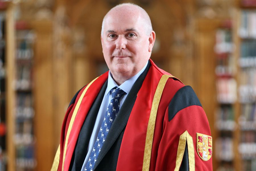 Man with close cropped hair stands in red graduation gown in Bangor University's library