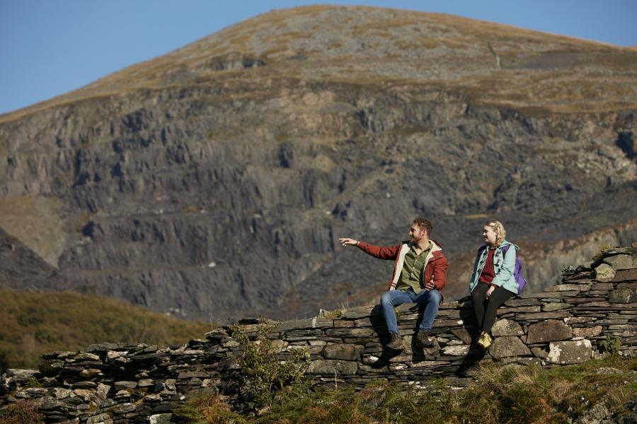 Young aman and woman sit on a slate wall, in the background is an enormous slate quarry in the side of a mountain