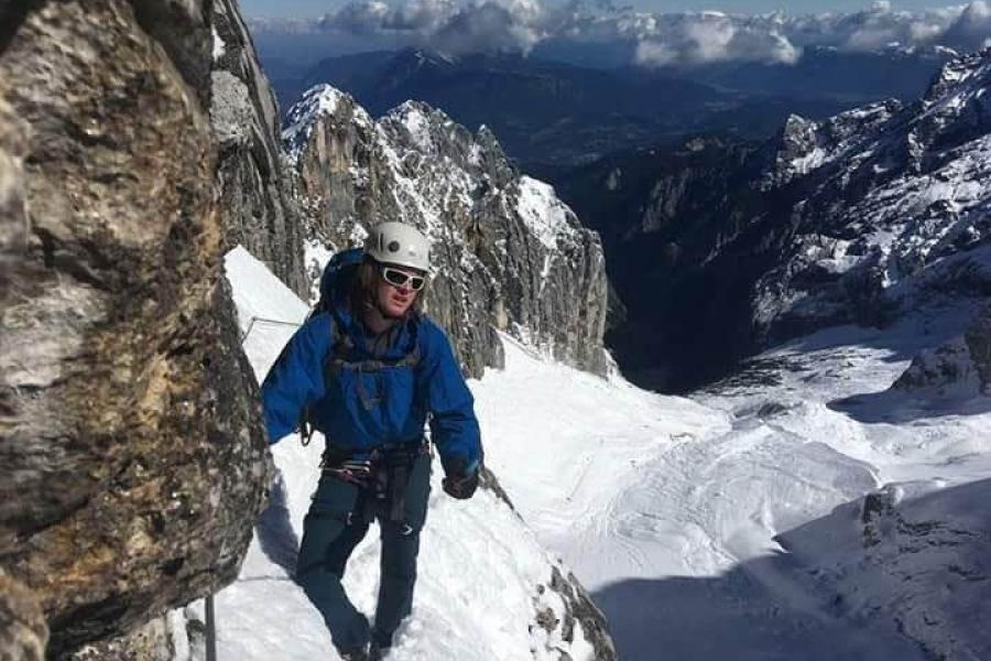 A man in Alpine climbing kit with helmet and sunglasses at the side of a snowy rocky precipice, on a rocky outcrop/