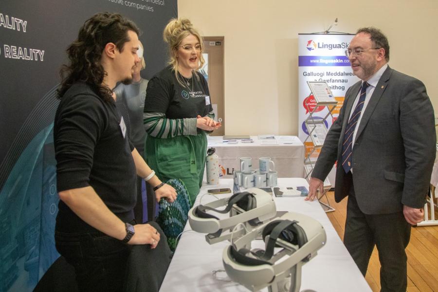  Dau berson ifanc ynsiarad ardraws bwrdd efo'r Is-ganghellor sy'n gwisgo siwt. Ym mlaen y llun mae dau penwisg rhithwirss a table with Vice Chancellor in a suit. There are VR headsets in the foreground