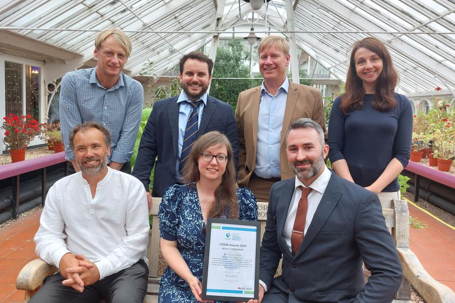 seven people two women five men in a greenhouse with three seated on a bench and four behind,    a woman in the centre of the bench holds a certificate.