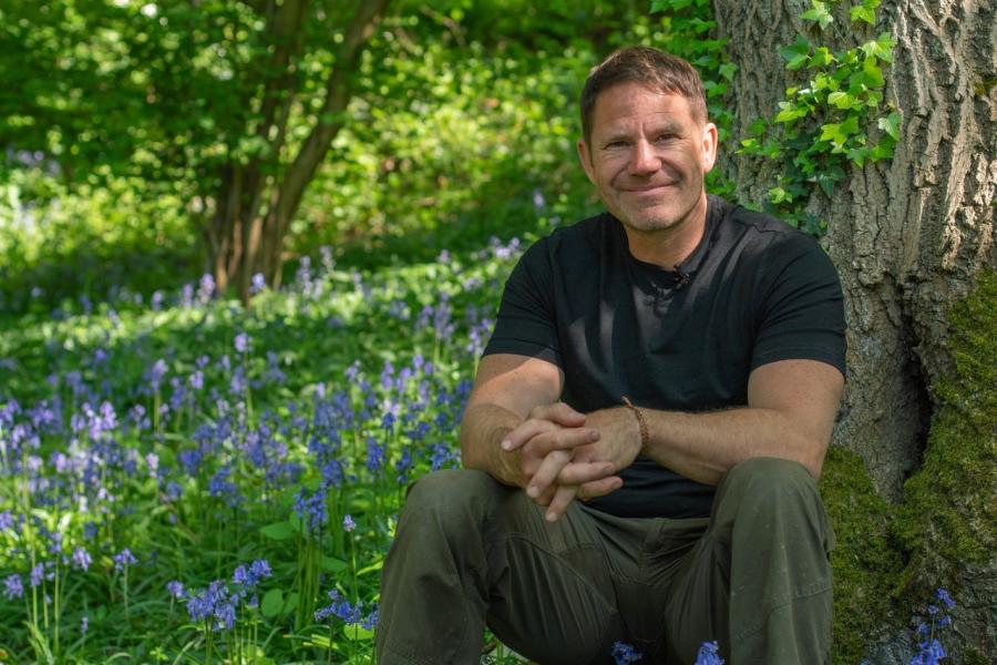 Eisteddai Steve Backshall tu allan, mae clychau'r gog mewn blodau o'i gwmpas a choed yn y cefndir.