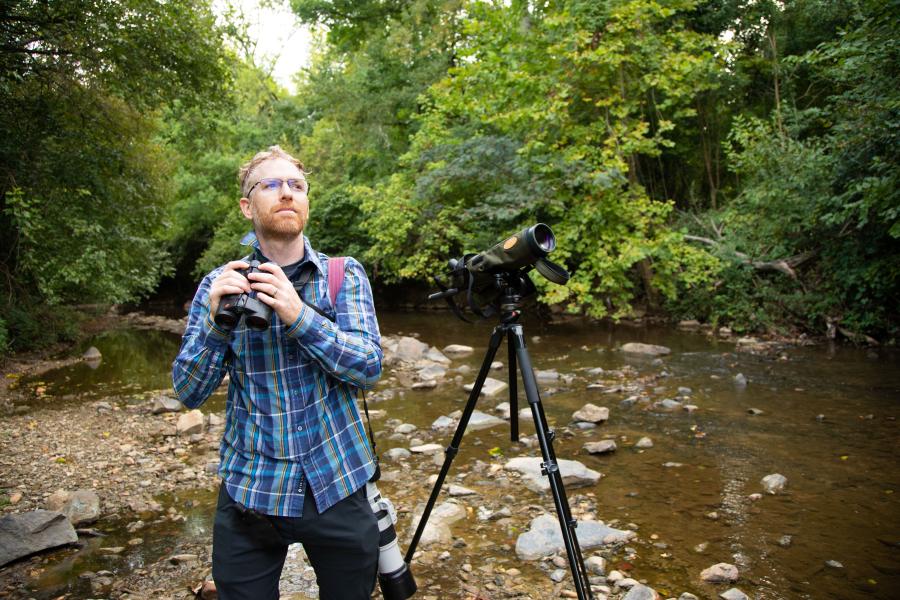 Dr Tyler Hallman standing in a stream surrounded by trees with a scope and binoculars in his hands