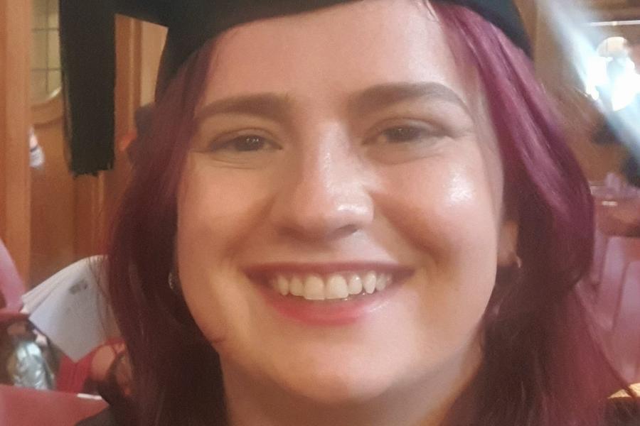 Portrait image of woman with purple hair smiling in Bangor University graduation cap and robes