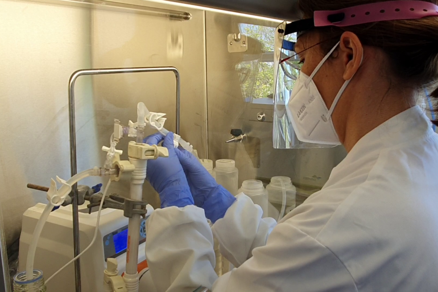 The image shows a person working in a laboratory setting. The individual is wearing protective gear, including a white lab coat, a face mask, gloves, and a face shield with a pink headband. They are handling laboratory equipment inside a fume hood or a biosafety cabinet, which is likely being used to ensure a sterile and safe environment for experiments. Various tubes, containers, and scientific instruments are visible, suggesting that the person is conducting a precise scientific procedure or experiment. T