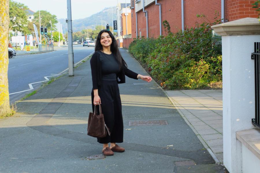 Shreyasi (Cece) Desai in black top and black skirt standing on a pavement holding a brown bag with the street behind