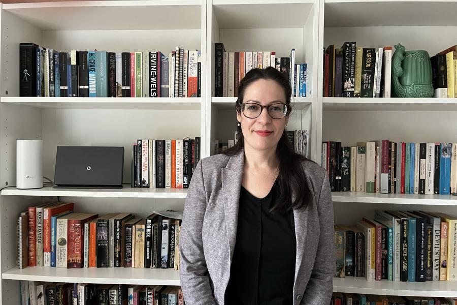 Dr Beverly Pickard-Jones wearing glasses and black top and blazer standing in front of a bookshelf