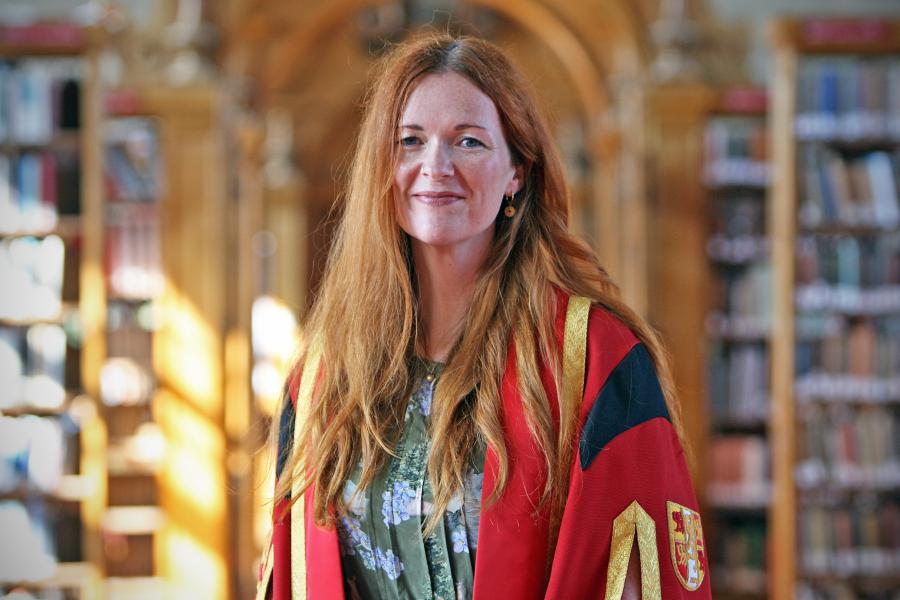 A female wearing wearing red and gold graduation robe with bookshelves in background