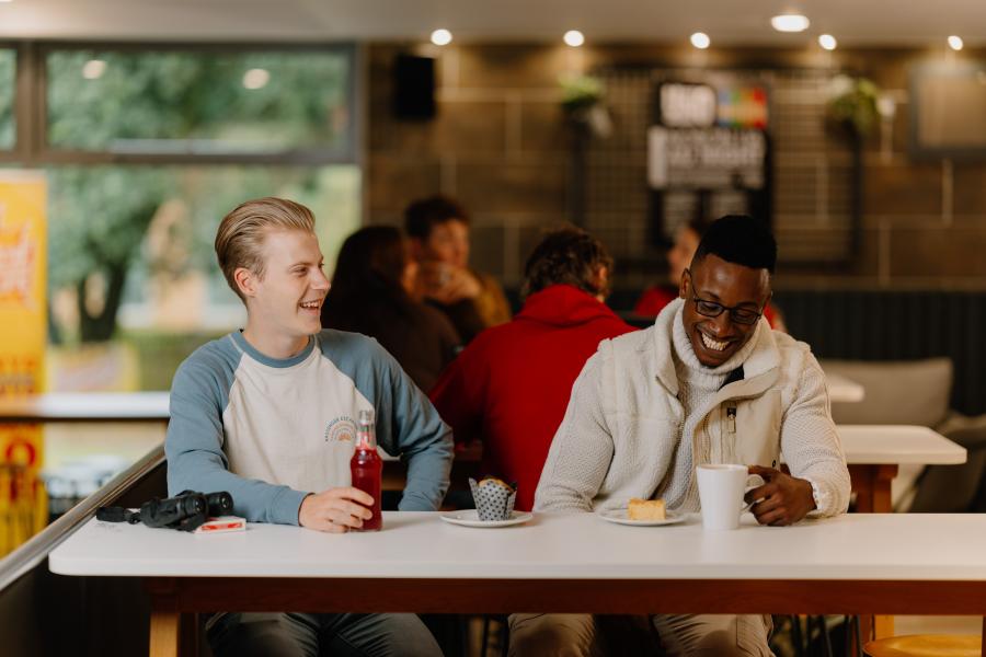 Two students sit laughing in Bar Uno, surrounded by groups of other students chatting and relaxing in the laid-back atmosphere.