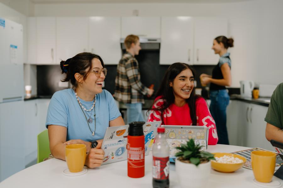 Two students sitting, chatting and laughing around a table in one of St Mary's  Village's shared kitchen while two other students are preparing food in the background.