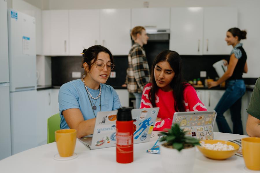 Four students in a shared kitchen in St Mary's halls. Two are sitting by the table looking at a laptop and two are chatting by the kitchen counter in the background.