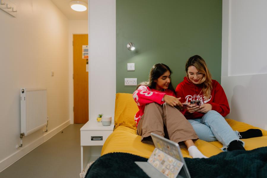 Two students sitting on a bed looking at a moblie phone in a room in St Mary's halls. 