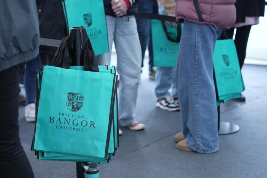 A queue of prospective students for the Open Day, the shot shows their legs and a pile of Open Day tote bags.