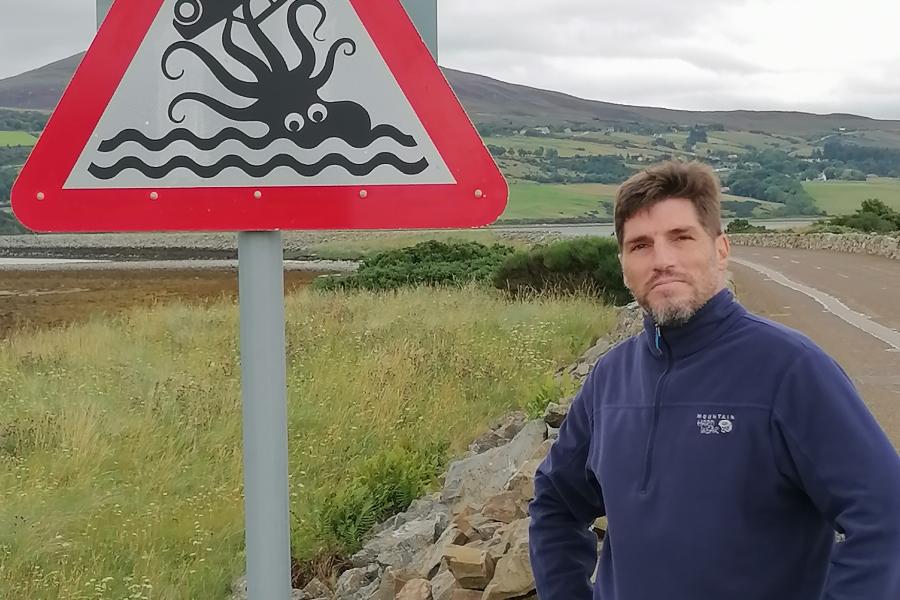 Tommie standing by a red black and white traffic sign displaying an octopus and a car with green scenery behind him. He's wearing a blue fleece.