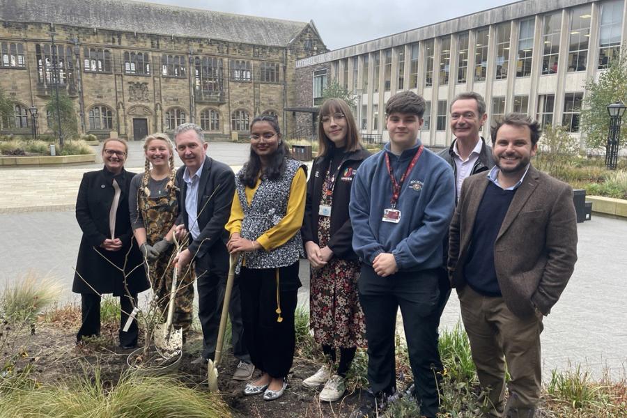 A group of men and women standing by a newly planted tree with university buildings in the background