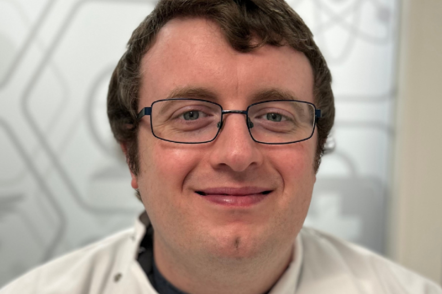 a headshot of Dr Dylan Jones, a lecturer with short brown hair and glasses wears a labcoat in front of a background of scientific symbols