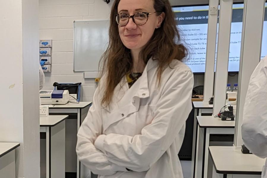 A female lecturer wearing a lab coat with long brown hair is stood in a laboratory