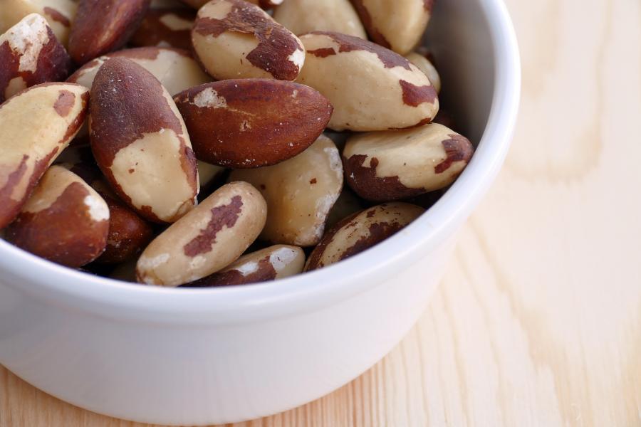 A white bowl containing shelled brazil nuts is in the left of the image, on a wooden table background